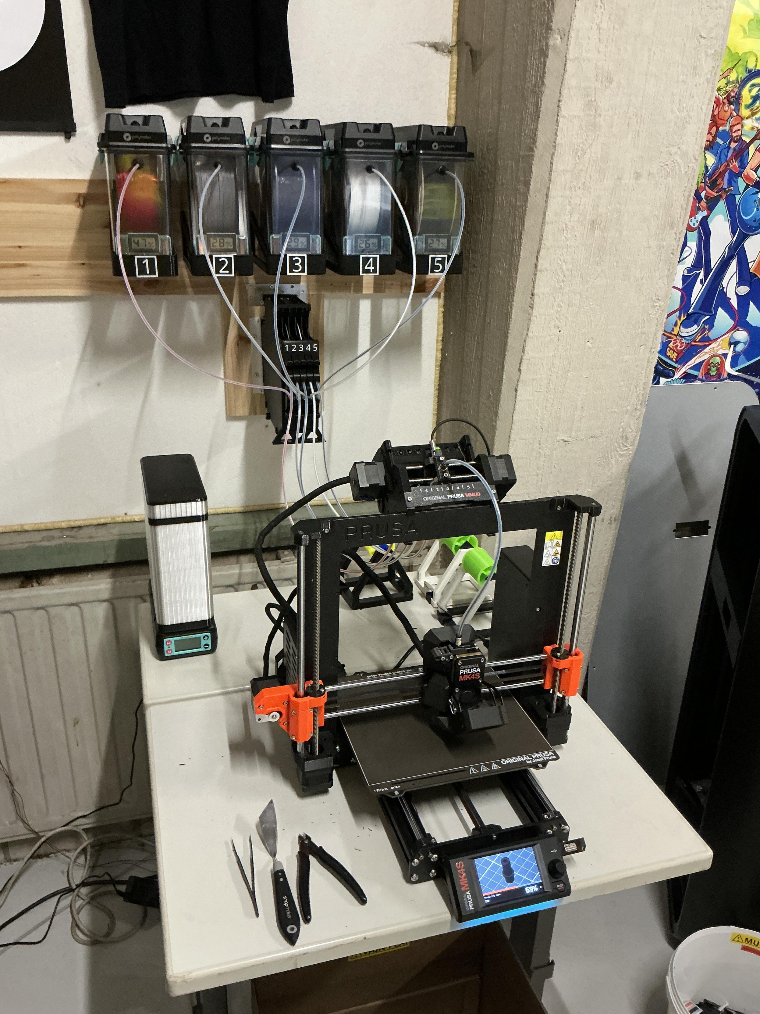 A Prusa Mk4S on a desk, with the dry boxes behind it on the wall.